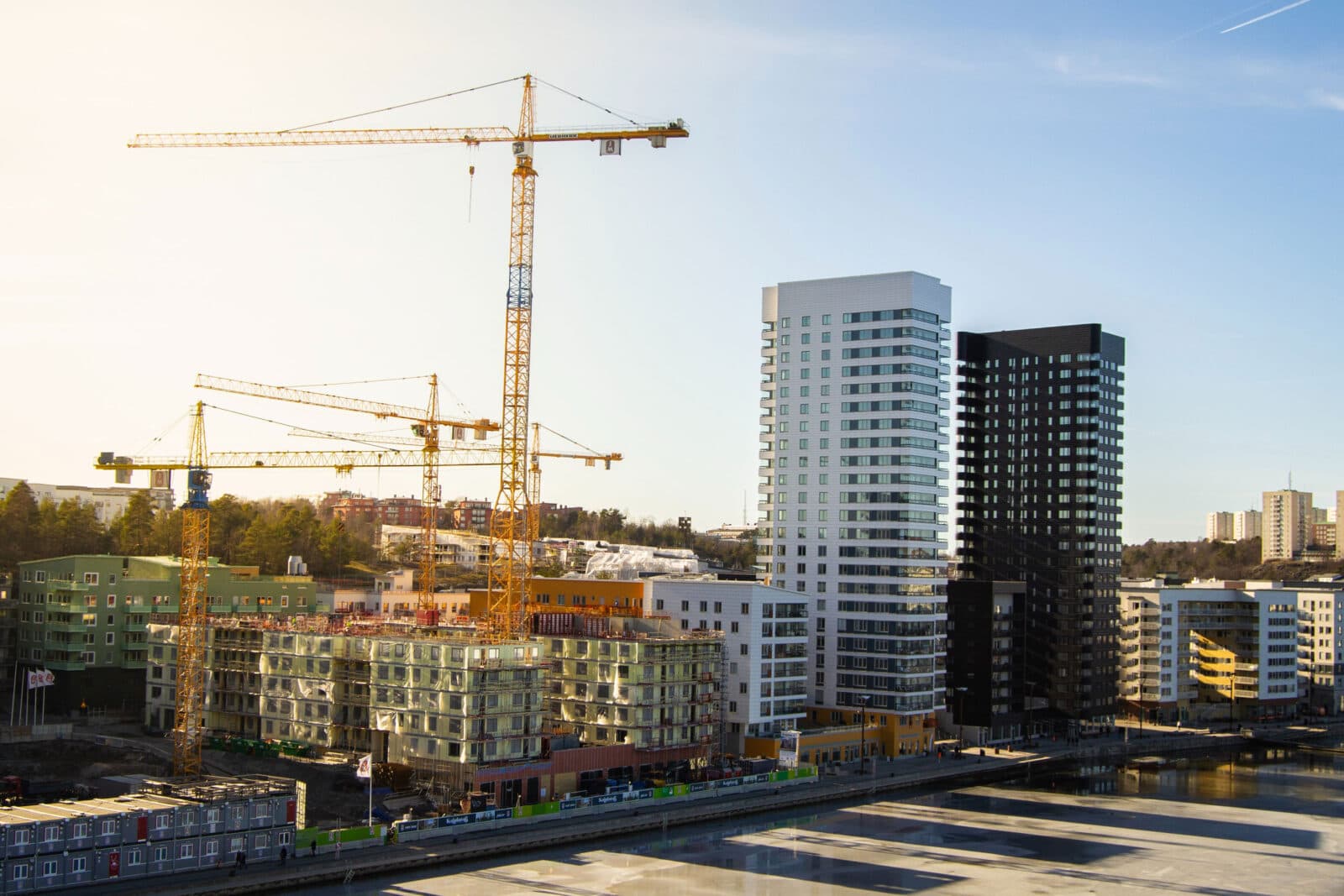 A construction site with cranes and tall buildings in the background. The cranes are tall and have long arms that extend over the construction site. The buildings in the background are tall and made of concrete and glass. There is a lot of activity on the construction site, with workers moving around and machinery operating.