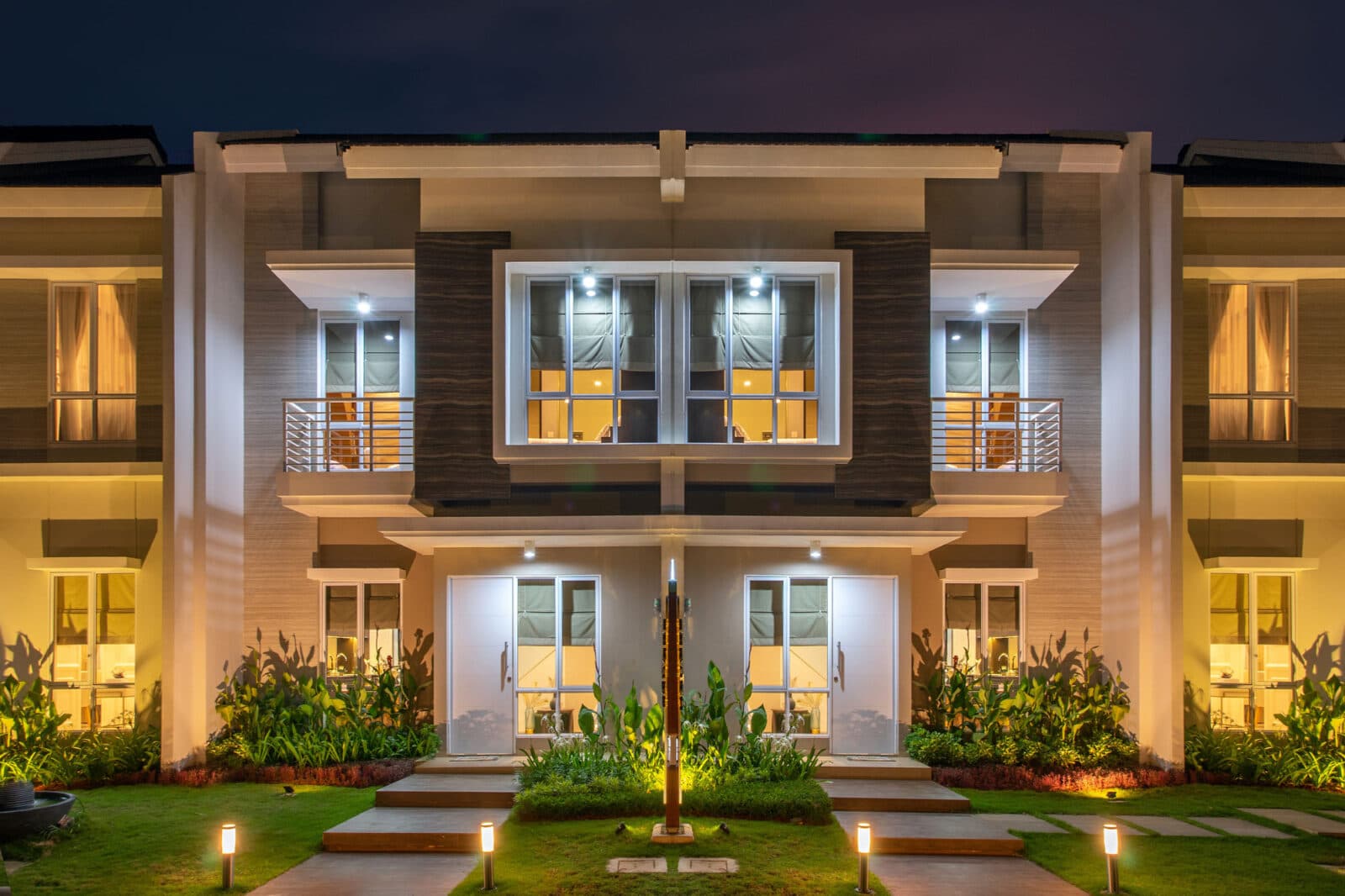 A two-floor duplex building in a row of such, in a modern style with many windows, balconies, and lawns with cactuses and shrubbery.